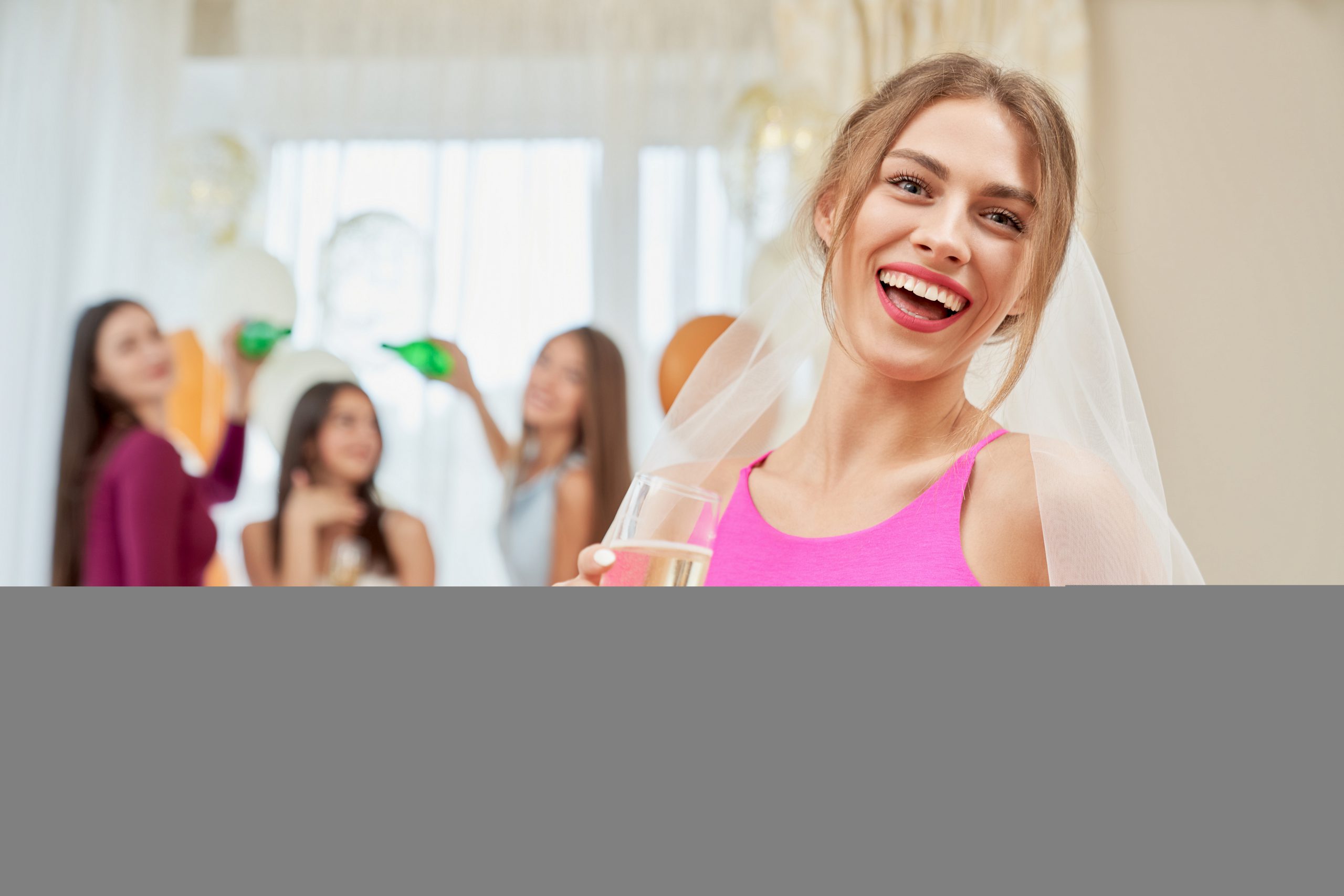 Uśmiechnięta panna młoda w welonie i różowym topie wznosi toast podczas imprezy. W tle rozmyte przyjaciółki i balony, tworząc atmosferę na wieczór panieński w Katowicach.