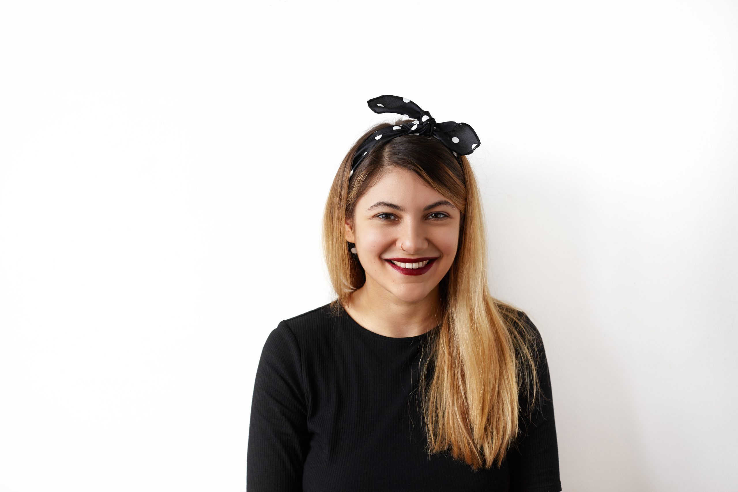 Uśmiechnięta kobieta w czarnej opasce z kokardą w białe grochy, która jest elementem fryzury podobnym do gumki z kokardą