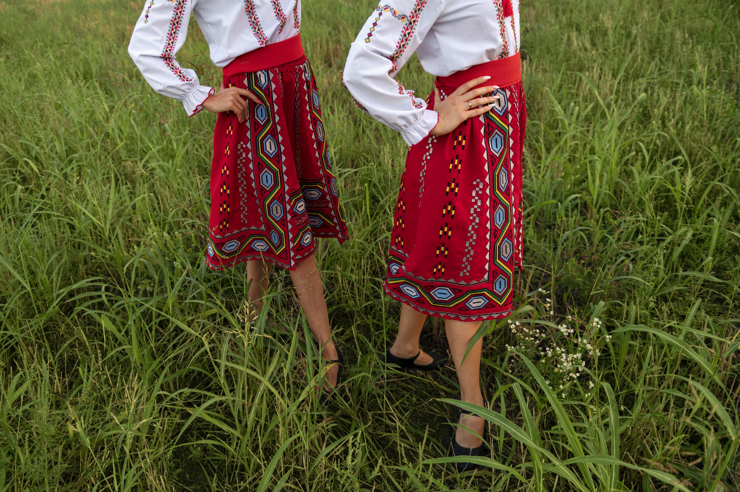 Dwie kobiety ubrane w czerwone spódnice folk z geometrycznym haftem, stojące bokiem w wysokiej, zielonej trawie.