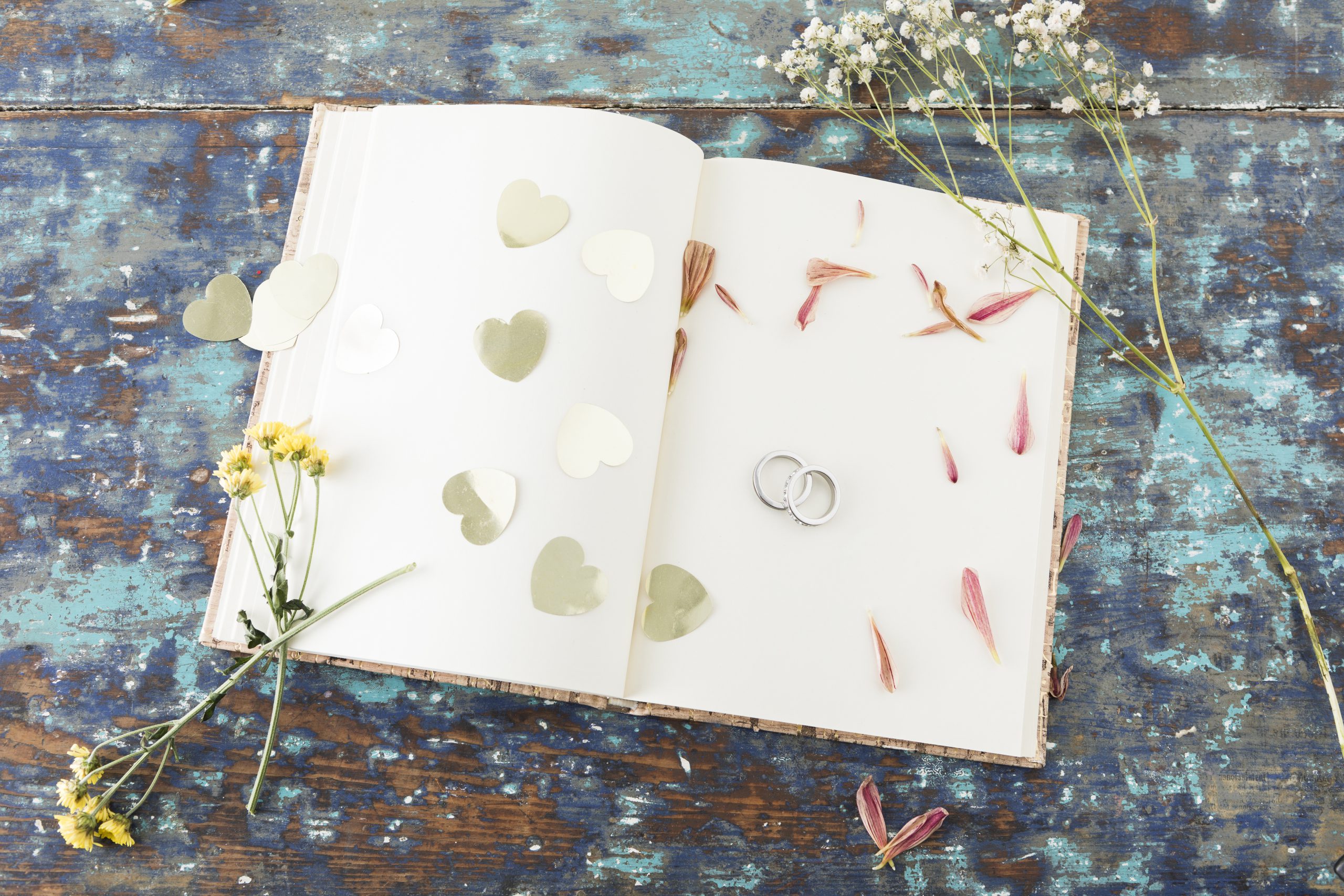 Otwarta księga gości w stylu boho, leżąca na starym, niebieskim drewnianym stole, obsypana konfetti w kształcie serc, płatkami kwiatów, gipsówką oraz obrączkami.
