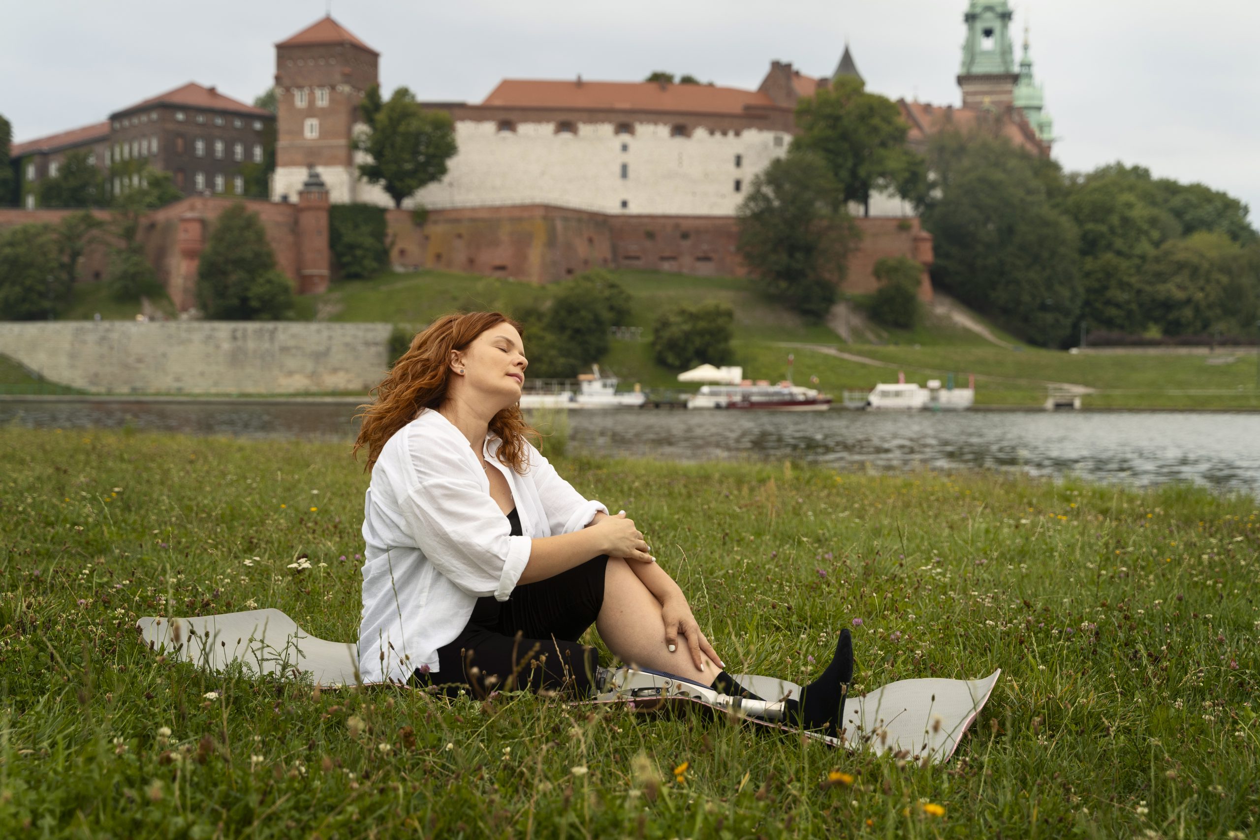 Kobieta z rudymi włosami i protezą nogi odpoczywa na macie na Bulwarach Wiślanych, w tle widoczny jest Zamek Królewski na Wawelu, stanowiący jedną z głównych attractions cracow.